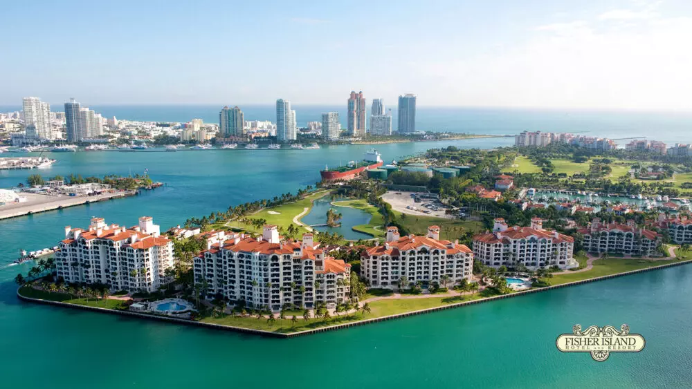 Aerial view of Fisher Island, showing luxury condominiums, greenery, and water surrounded by high-rise buildings and the ocean in the background.