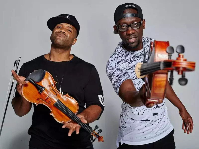 Two men holding black violins and wearing casual clothes and hats pose confidently against a plain background.