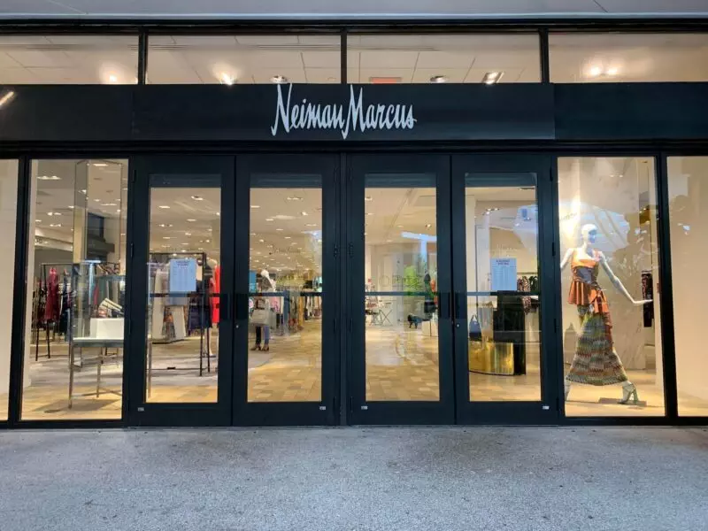 Exterior of a Neiman Marcus store with large glass doors. The interior features clothing displays and mannequins.