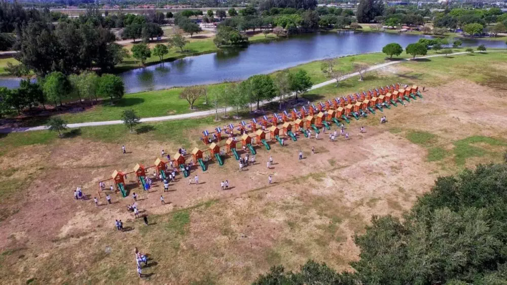 Aerial view of a large outdoor playground featuring the longest swing set alongside multiple colorful play structures in a park setting. A lake and trees are visible in the background, with people scattered throughout the area.