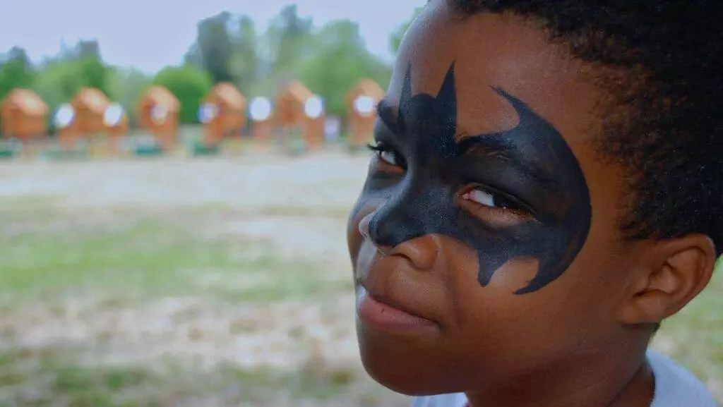 A child with Batman face paint looks at the camera with a slight smile. The background shows a blurry outdoor setting, hinting at wooden structures, possibly near the longest swing set in town.