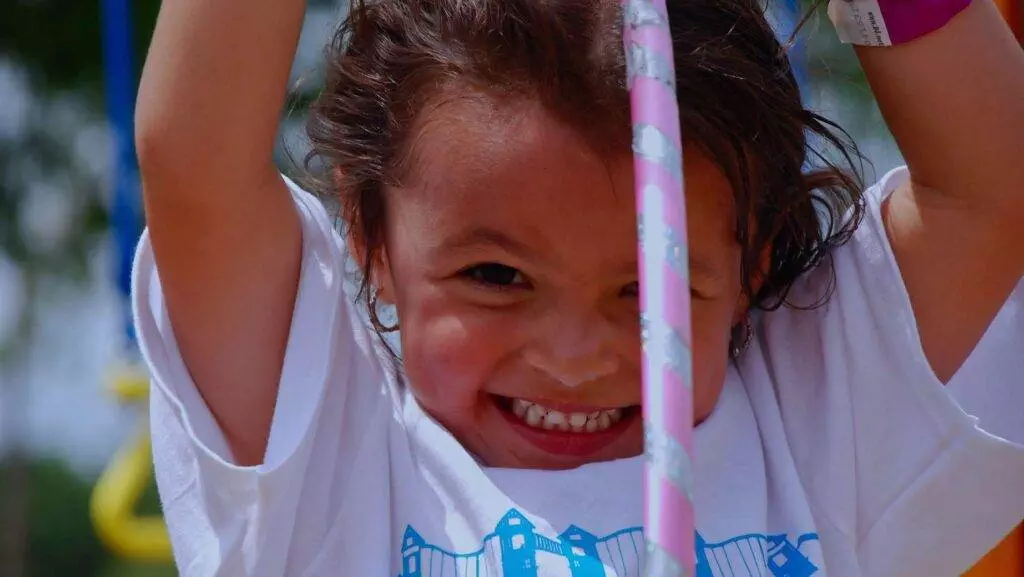 A child with a white shirt smiles brightly while holding onto a pink-striped rope with both hands, enjoying the longest swing set outdoors.