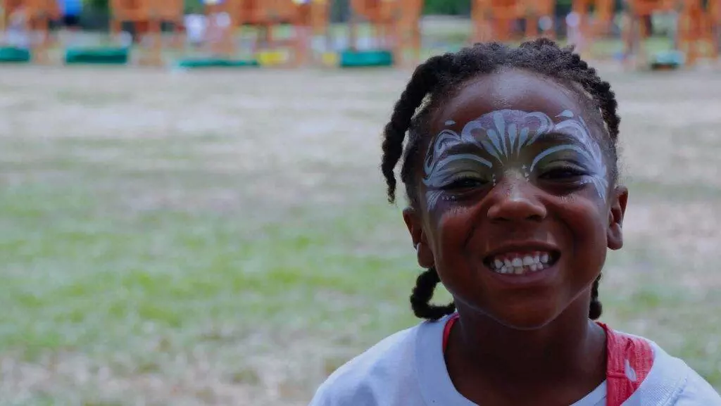 A child with face paint smiles broadly while outdoors, with the longest swing set visible in the blurred playground background.