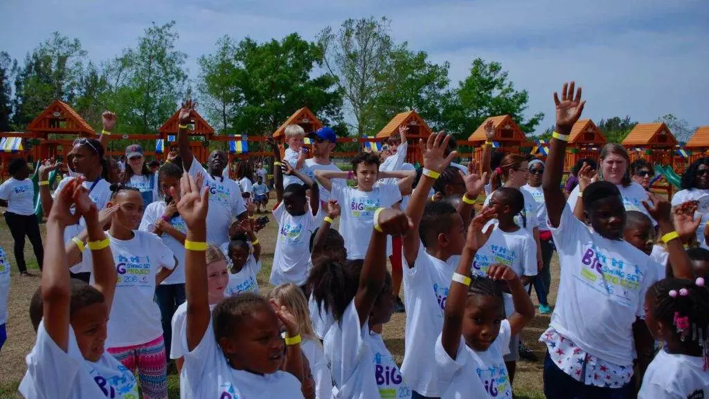 A large group of children and adults wearing matching white T-shirts participate in an outdoor event, raising their hands in excitement. Wooden structures and trees are visible in the background, possibly surrounding the longest swing set.