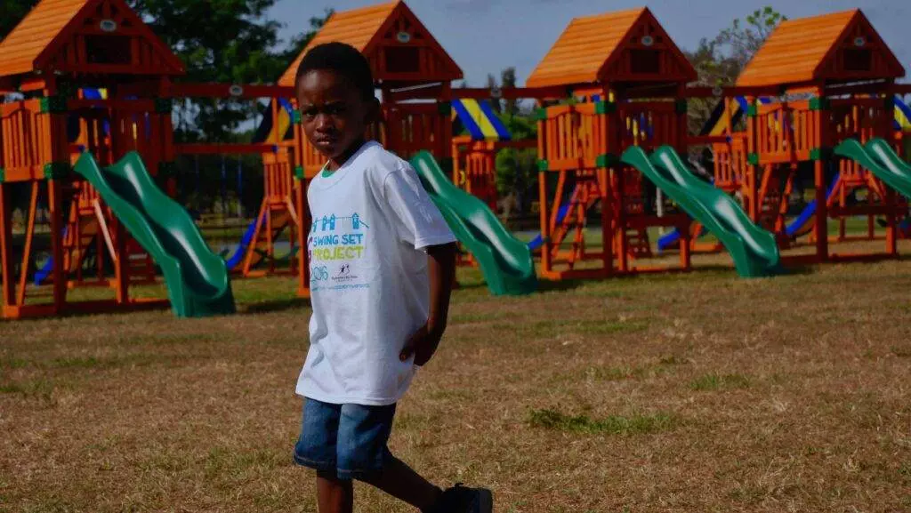 A young child wearing a white T-shirt and denim shorts stands on a grassy field in front of a playground with multiple green slides, orange structures, and the longest swing set in the park.