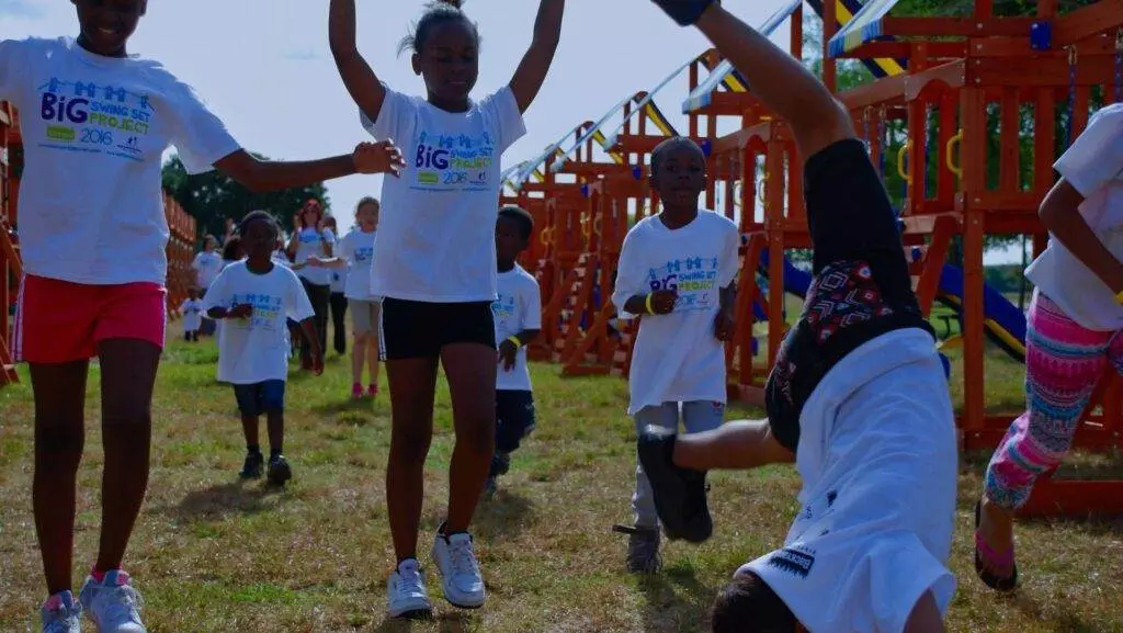 Children in matching T-shirts play on a playground, their laughter filling the air. One child does a cartwheel while others run and jump around, eyeing the longest swing set with excitement.