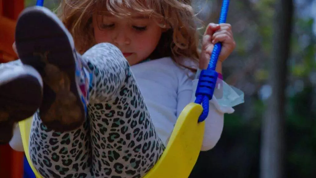 A child with blonde hair is swinging on a yellow swing, part of the longest swing set in the park. The child is wearing a white shirt and leopard print pants, and the background shows blurred outdoor scenery.