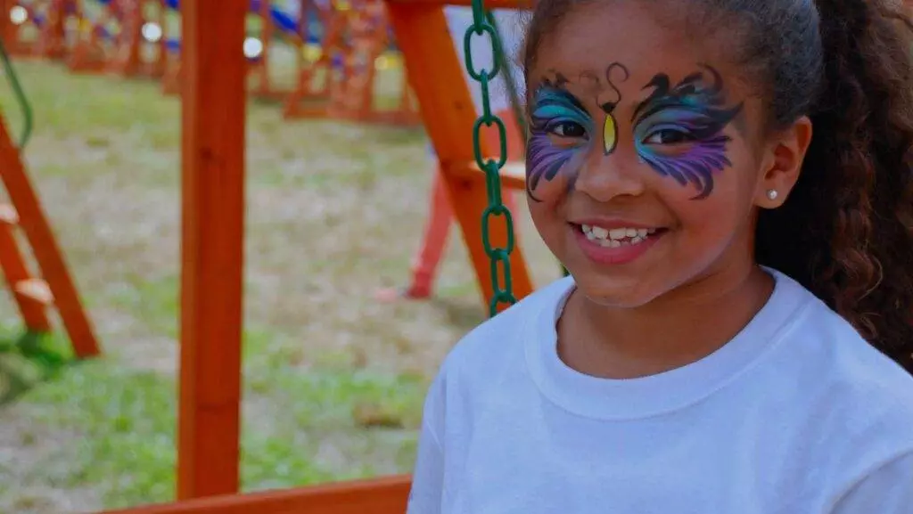 A young girl with colorful face paint smiles while sitting on a swing in the playground’s longest swing set.