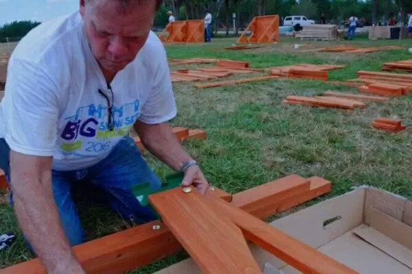 A man wearing a white t-shirt assembles wooden components on the grass at an outdoor event, preparing what appears to be the longest swing set ever built.